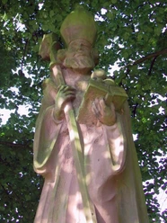 Das Nikolausdenkmal. Steinernes Nikolausdenkmal unter grünem Laub, zeigt Nikolaus mit Bischofsstab und Buch.