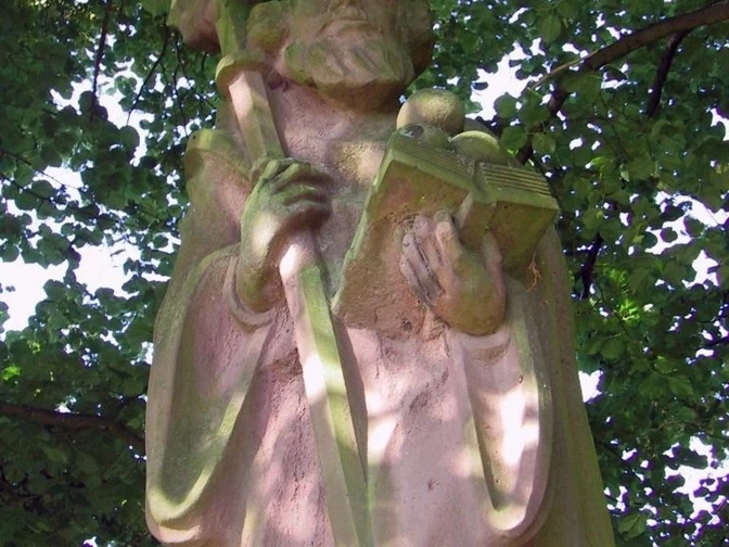 Das Nikolausdenkmal. Steinernes Nikolausdenkmal unter grünem Laub, zeigt Nikolaus mit Bischofsstab und Buch.