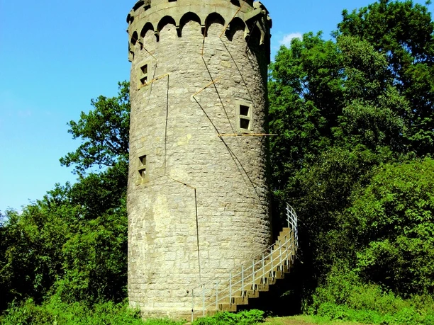 Auf unserer Wanderung kommen wir am Holsterturm vorbei. Ein historischer Steinturm mit Wendeltreppe steht von Bäumen umgeben im Grünen unter klarem Himmel.
