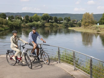 Zwei Radfahrer auf einer Brücke über einen ruhigen Fluss, umgeben von grüner Landschaft.