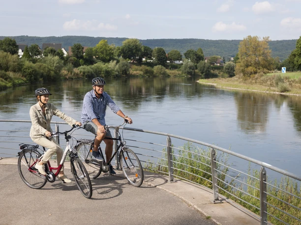 Werre-Weser-Kuss Zwei Radfahrer auf einer Brücke über einen ruhigen Fluss, umgeben von grüner Landschaft.