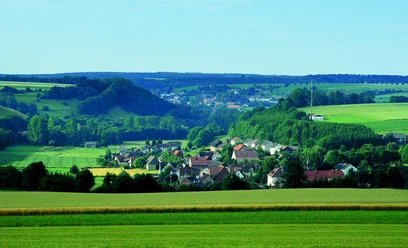 image Ein kleines Dorf eingebettet in eine grüne Hügellandschaft mit weitem Blick auf Felder und Wälder.