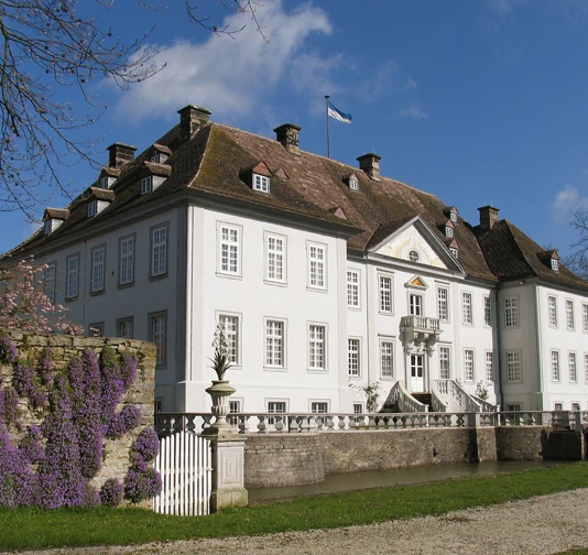 Wasserschloß Vinsebeck Weißes Wasserschloss mit vielen Fenstern, umgeben von lila Blumen und alten Mauern unter blauem Himmel.