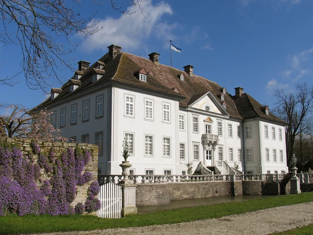 Wasserschloß Vinsebeck Weißes Wasserschloss mit vielen Fenstern, umgeben von lila Blumen und alten Mauern unter blauem Himmel.