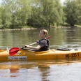 Person in einem orangefarbenen Kanu paddelt auf einem ruhigen Fluss, umgeben von grüner Natur.