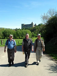 image Drei Wanderer in Freizeitkleidung auf einem Weg, im Hintergrund ein großes Schloss auf einem Hügel.