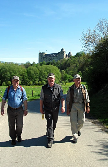 image Drei Wanderer in Freizeitkleidung auf einem Weg, im Hintergrund ein großes Schloss auf einem Hügel.