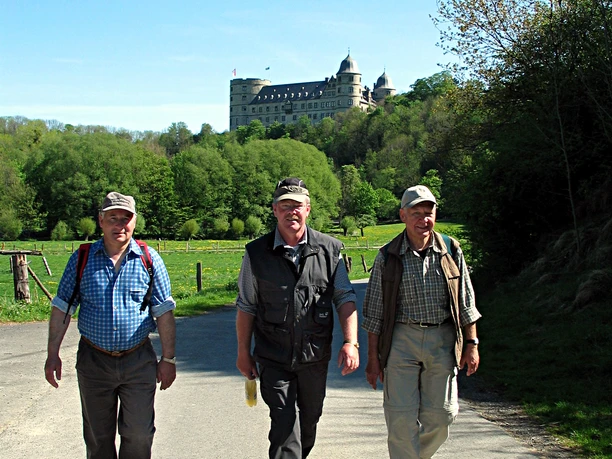 image Drei Wanderer in Freizeitkleidung auf einem Weg, im Hintergrund ein großes Schloss auf einem Hügel.