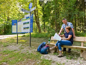 Wanderer am Wanderinfopunkt Ein Paar sitzend auf einer Holzbank vor einem Wegweiser im sonnigen Wald, Karten studierend.