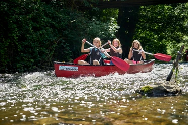 Kanutour Bega und Werre Drei Personen paddeln in einem roten Kanu auf einem flachen, klaren Fluss unter sonnigem Himmel.