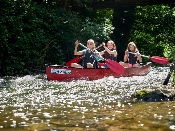 Kanutour Bega und Werre Drei Personen paddeln in einem roten Kanu auf einem flachen, klaren Fluss unter sonnigem Himmel.