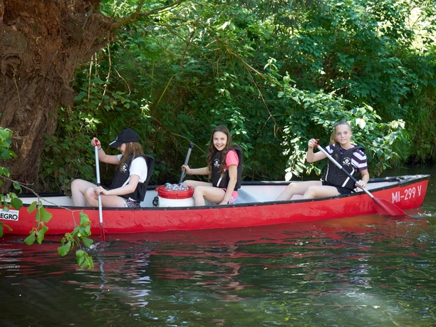 Kanutour Bega und Werre Drei junge Menschen paddeln fröhlich in einem roten Kanu auf einem grünen Fluss umgeben von Bäumen.