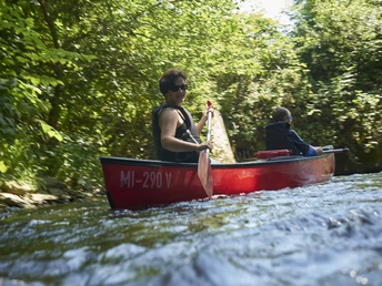 Kanutour Bega und Werre Kanu auf sprudelndem Fluss, zwei Personen paddeln im roten Boot, umgeben von dichter Vegetation.