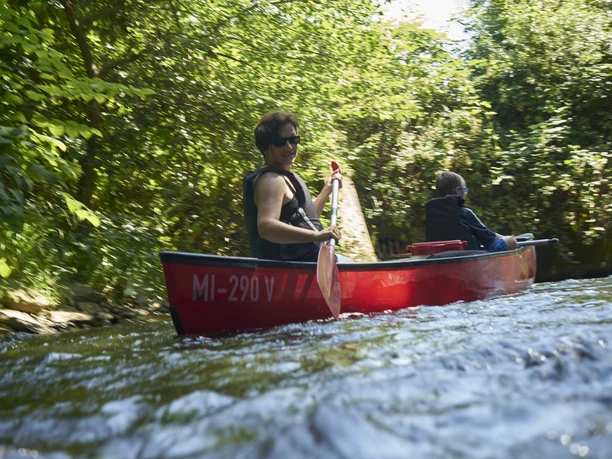 Kanutour Bega und Werre Kanu auf sprudelndem Fluss, zwei Personen paddeln im roten Boot, umgeben von dichter Vegetation.