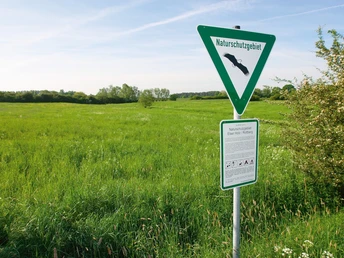 Grüne Wiesen und ein Schild mit Hinweis auf ein Naturschutzgebiet unter blauem Himmel.