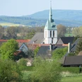 Bergheimer Kiche Weißer Kirchturm mit grünem Dach, umgeben von Häusern und Bäumen, vor hügeliger Landschaft.