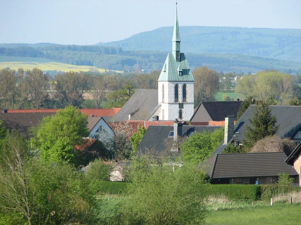 Bergheimer Kiche Weißer Kirchturm mit grünem Dach, umgeben von Häusern und Bäumen, vor hügeliger Landschaft.