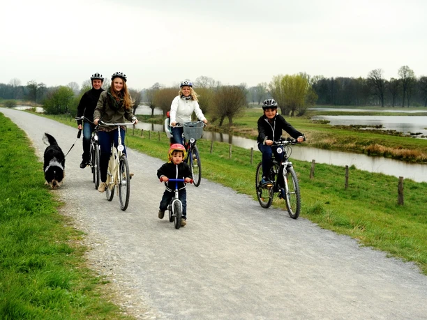 Radler am Steinhorster Becken Radfahrer mit Hund und Kind auf einem Schotterweg entlang eines ruhigen Gewässers in ländlicher Umgebung.