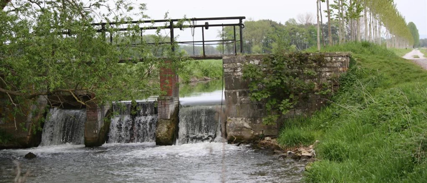 Schleuse am Boker Kanal Steinernes Bauwerk mit Schleuse über ruhigem Fluss, von Bäumen gesäumte Allee im Hintergrund.