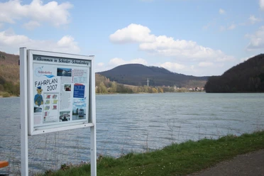 Schiedersee Ein See mit bewaldeten Hügeln im Hintergrund, ein Schild mit Informationen am Ufer, blauer Himmel.
