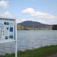 Schiedersee Ein See mit bewaldeten Hügeln im Hintergrund, ein Schild mit Informationen am Ufer, blauer Himmel.