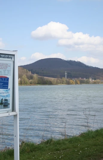 Schiedersee Ein See mit bewaldeten Hügeln im Hintergrund, ein Schild mit Informationen am Ufer, blauer Himmel.