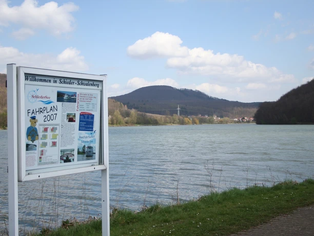 Schiedersee Ein See mit bewaldeten Hügeln im Hintergrund, ein Schild mit Informationen am Ufer, blauer Himmel.