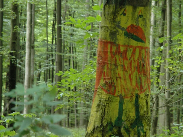 Kunst im Wald Ein Baum im Wald, dekoriert mit bunter, kindlicher Malerei eines stilisierten Tieres.