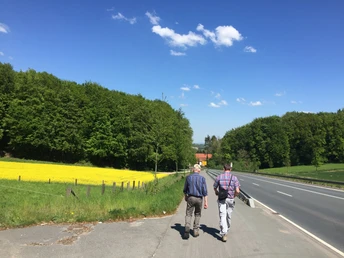 Zwei Wanderer auf dem Rundwanderweg Zwei Männer spazieren auf einem ländlichen Weg neben einer Straße, umgeben von grünen Wäldern und einem blühenden Feld.