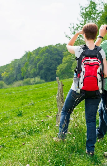 Ein Paar mit Wanderrucksäcken blickt von einer grünen Wiese aus mit Ferngläsern in die Ferne.