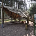 Ein hölzerner Unterstand im Wald, mit Sitzbänken und Informationstafel, umgeben von hohen Bäumen.