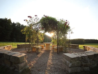 Eine steinerne Sitzgruppe umgeben von einem Rosengitter, beleuchtet von der tief stehenden Sonne.