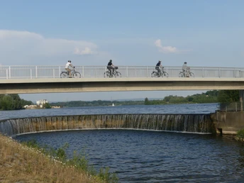 Fußgänger und Radfahrer auf einer Brücke überqueren ein Auslaufbauwerk an einem Fluss bei Sonnenschein.