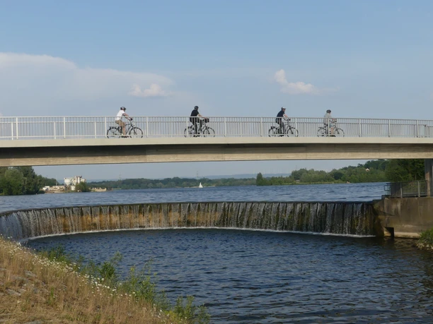 Auslaufbauwerk und Fußgänger-/Radfahrerbrücke Fußgänger und Radfahrer auf einer Brücke überqueren ein Auslaufbauwerk an einem Fluss bei Sonnenschein.