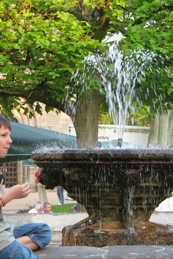 Steinheim, Kump Junge sitzt lächelnd auf einer Mauer neben einem plätschernden Brunnen umgeben von Grün.