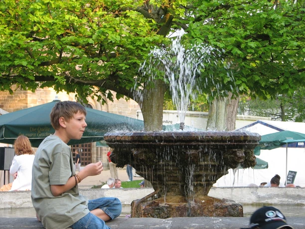 Steinheim, Kump Junge sitzt lächelnd auf einer Mauer neben einem plätschernden Brunnen umgeben von Grün.