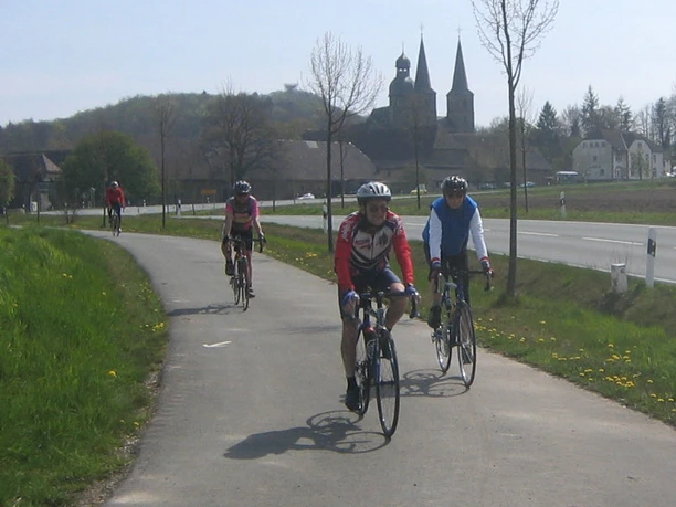 Vor der Abtei Marienmünster Radfahrer auf einem sonnigen Weg, im Hintergrund die markante Abtei Marienmünster und umliegende Felder.