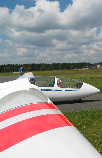 Ein Segelflugzeug am Boden, bereit zum Start, vor einer bewaldeten Landschaft und Wolken am Himmel.