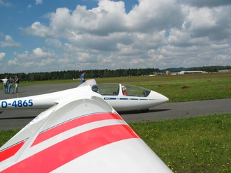 Segelflugplatz Ein Segelflugzeug am Boden, bereit zum Start, vor einer bewaldeten Landschaft und Wolken am Himmel.