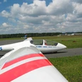 Segelflugplatz Ein Segelflugzeug am Boden, bereit zum Start, vor einer bewaldeten Landschaft und Wolken am Himmel.