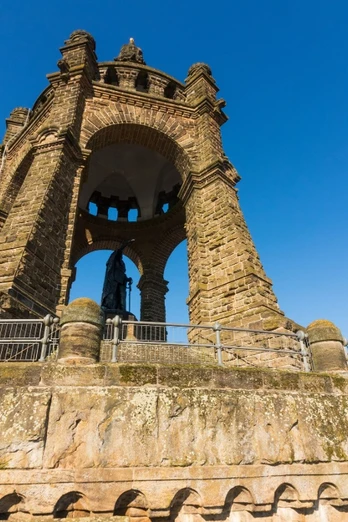 image Ein imposantes steinernes Denkmal mit offenen Bögen, eingefasst von Treppen, kontrastiert den blauen Himmel.