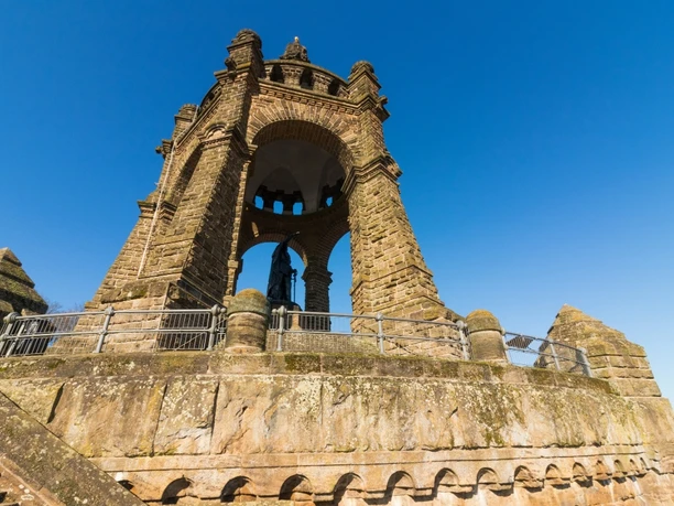 image Ein imposantes steinernes Denkmal mit offenen Bögen, eingefasst von Treppen, kontrastiert den blauen Himmel.