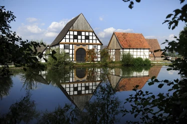 Der Dorfteich im LWL-Freilichtmuseum Detmold spiegelt Fachwerkhäuser und bewölkten Himmel wider.