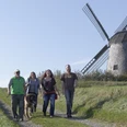 Windmühle Bavenhausen Vier Personen und ein Hund spazieren auf einem Pfad entlang einer historischen Windmühle bei sonnigem Wetter.