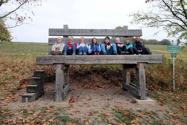 image Eine Gruppe von acht Personen sitzt fröhlich auf einer übergroßen Holzbank in herbstlicher Landschaft.