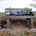 image Eine Gruppe von acht Personen sitzt fröhlich auf einer übergroßen Holzbank in herbstlicher Landschaft.