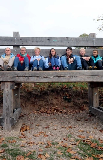 image Eine Gruppe von acht Personen sitzt fröhlich auf einer übergroßen Holzbank in herbstlicher Landschaft.