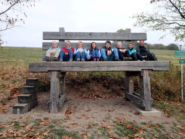 image Eine Gruppe von acht Personen sitzt fröhlich auf einer übergroßen Holzbank in herbstlicher Landschaft.