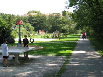Zwei Kinder spielen Tischtennis auf einem Parkweg, der von Bäumen und einem Spielplatz umgeben ist.