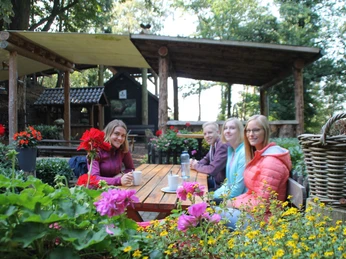 Rast an der Almhütter Vier Personen sitzen lächelnd an einem Holztisch im Freien, umgeben von bunten Blumen und Bäumen.